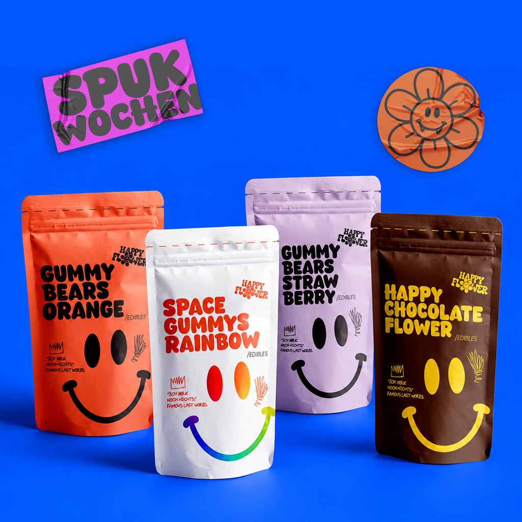 Vier Happy Flower Edibles-Baggys in Orange, Lila, Weiß und Braun, ästhetisch arrangiert auf leuchtend blauem Hintergrund. Die Verpackungen zeigen bunte Smiley-Designs und werfen natürliche, weiche Schatten.