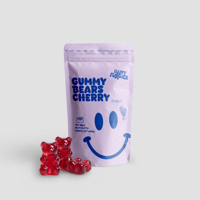Happy Flower Gummy Bears Cherry Edibles in lilafarbener Standbeutel-Verpackung mit mehreren roten Gummibärchen im Vordergrund auf hellem Hintergrund.